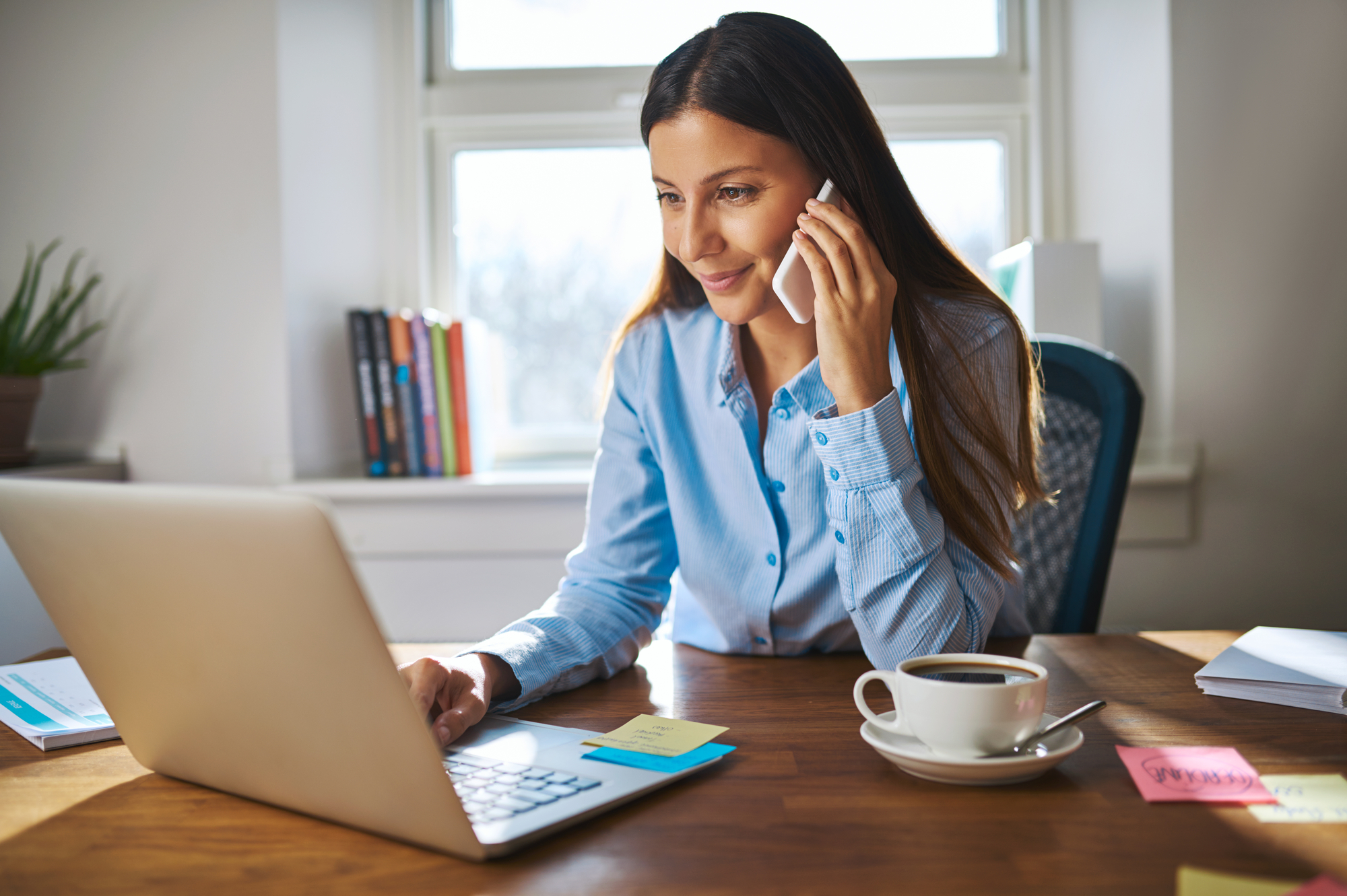 Young female adult on phone while working on laptop computer at desk next to coffee cup with large window in background