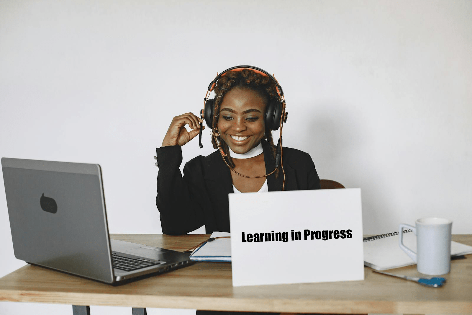 A smiling woman in headphones sits at a desk with a laptop, a notebook, and a mug, she engages in online learning, indicated by a sign saying "Learning in Progress."